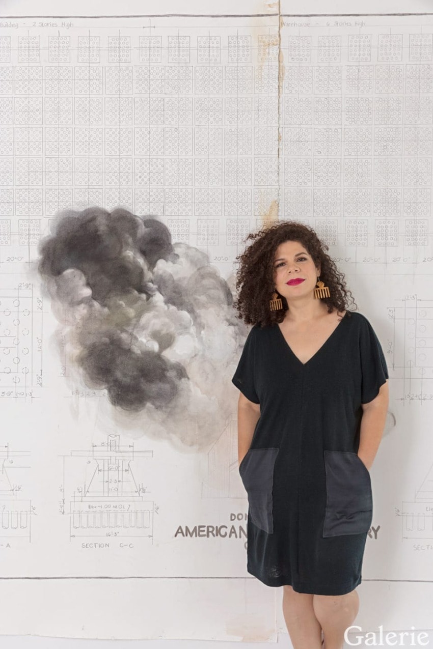 Person with curly hair wearing a black dress standing in front of a large artwork featuring clouds.