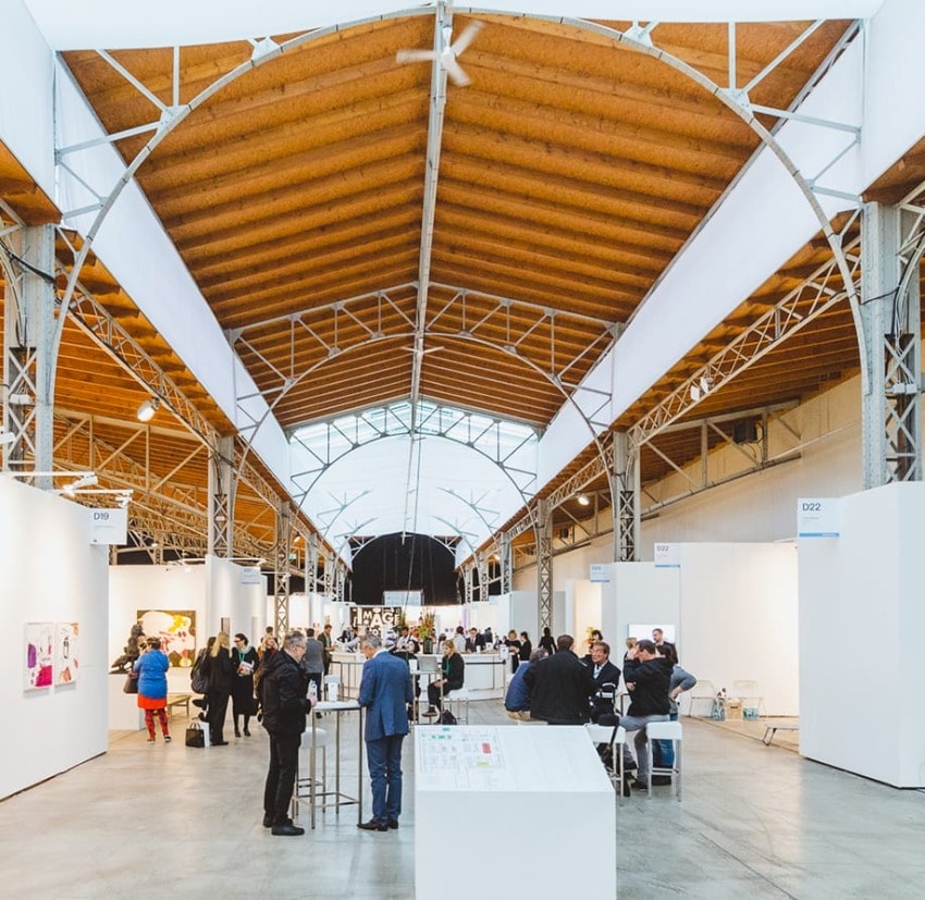 Art fair exhibition hall with people viewing and discussing artwork displayed in a spacious venue with a high wooden ceiling.