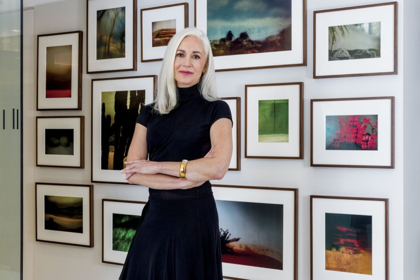 Person standing in front of a gallery wall with framed photographs, wearing a black outfit with folded arms.