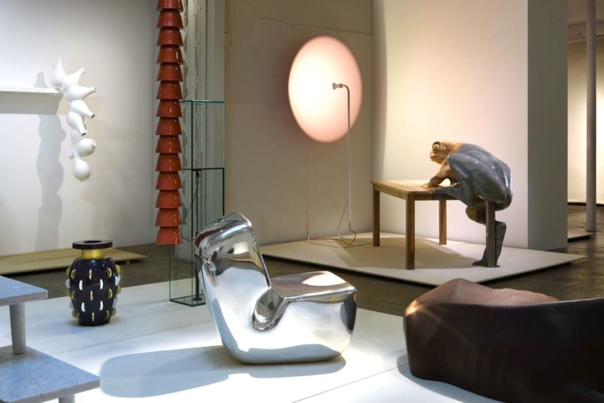 Modern art exhibition with abstract sculptures, a shiny chair, a stylized figure at a table, and various decorative pieces