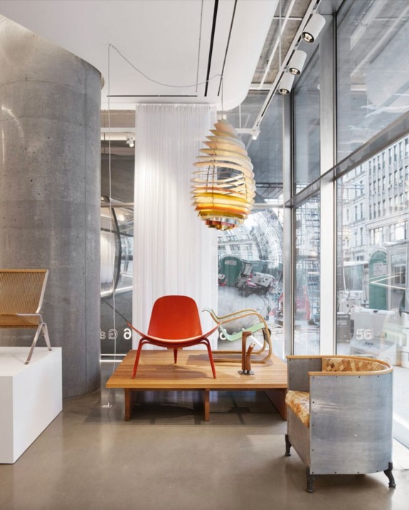 Modern interior design showroom with a red chair, a unique pendant light, a grey armchair, and large windows showcasing the city.