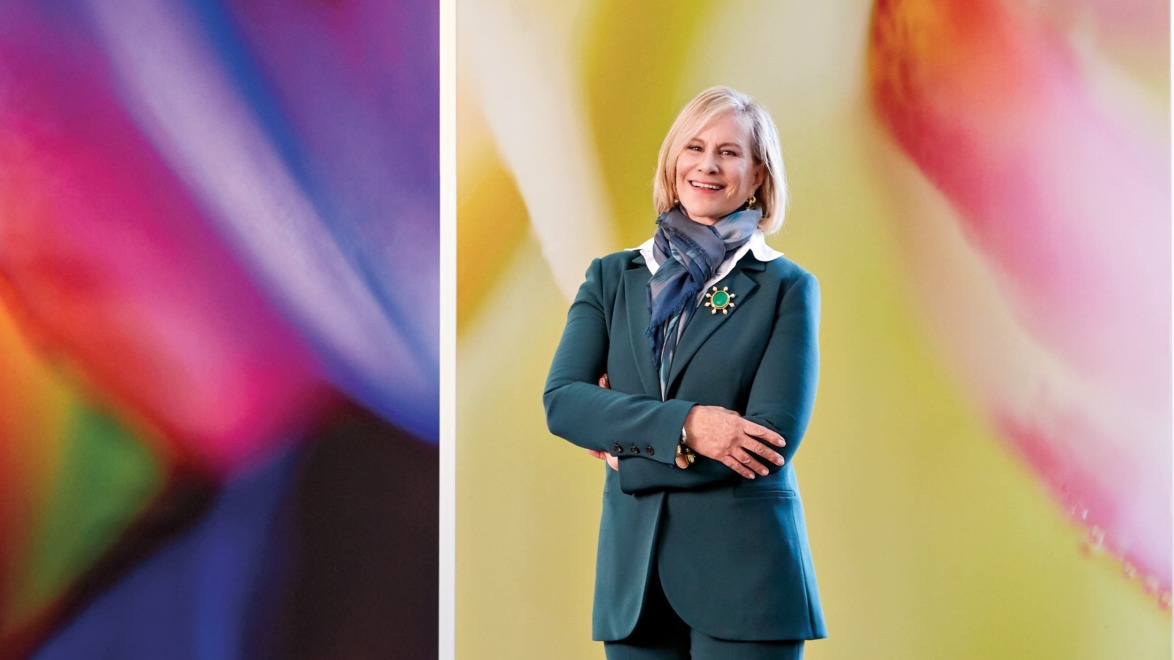Person in a teal suit and scarf standing in front of a colorful abstract background.