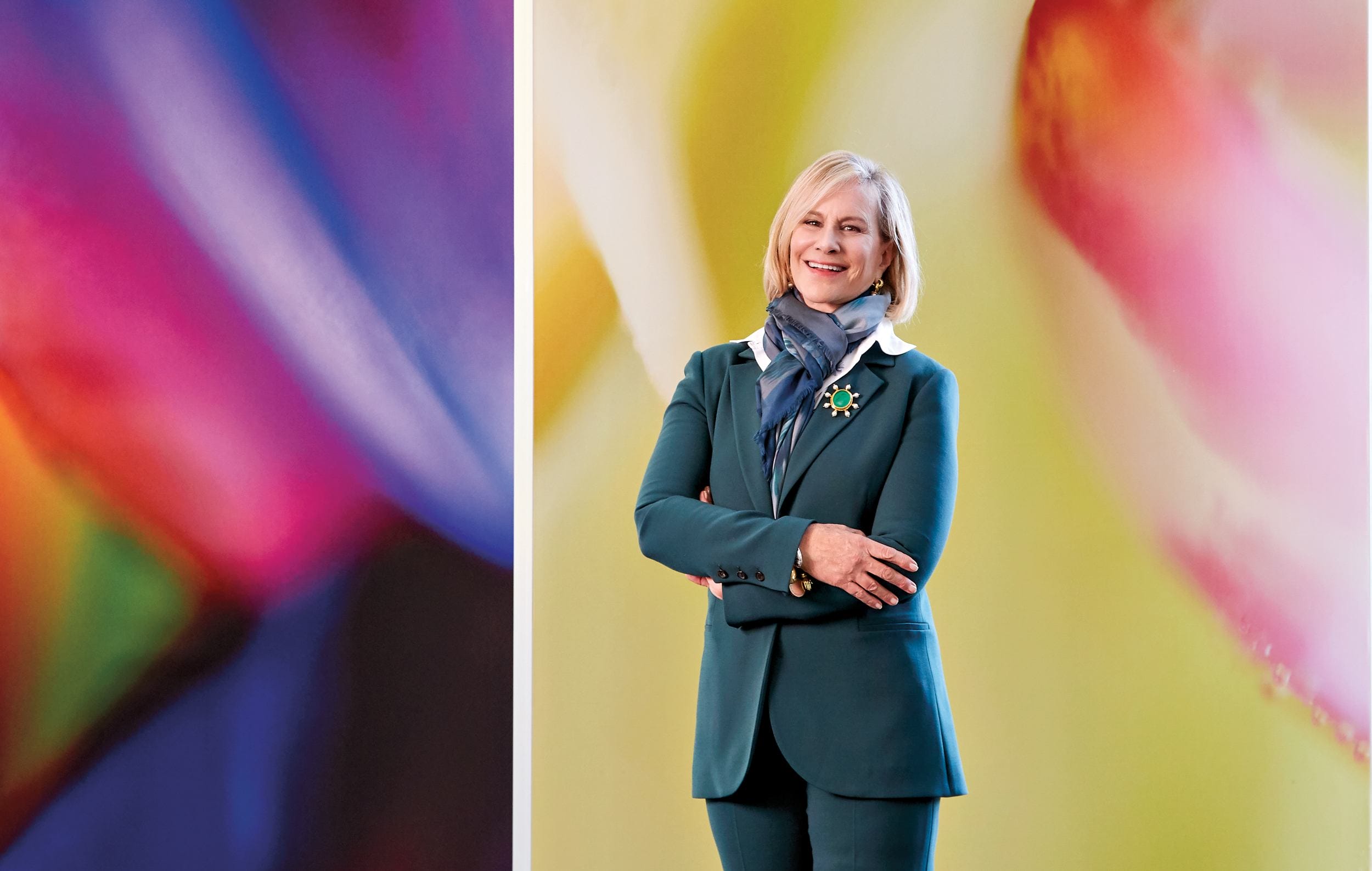 Person in a teal suit and scarf standing in front of a colorful abstract background.