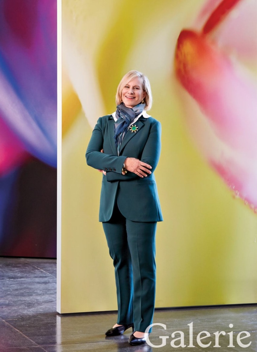 Person in a teal suit standing with arms crossed in a colorful art gallery setting.