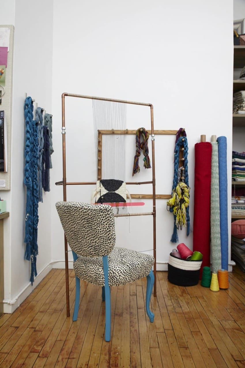Chair with leopard print fabric in front of weaving loom, surrounded by colorful yarns and fabrics on a wooden floor.