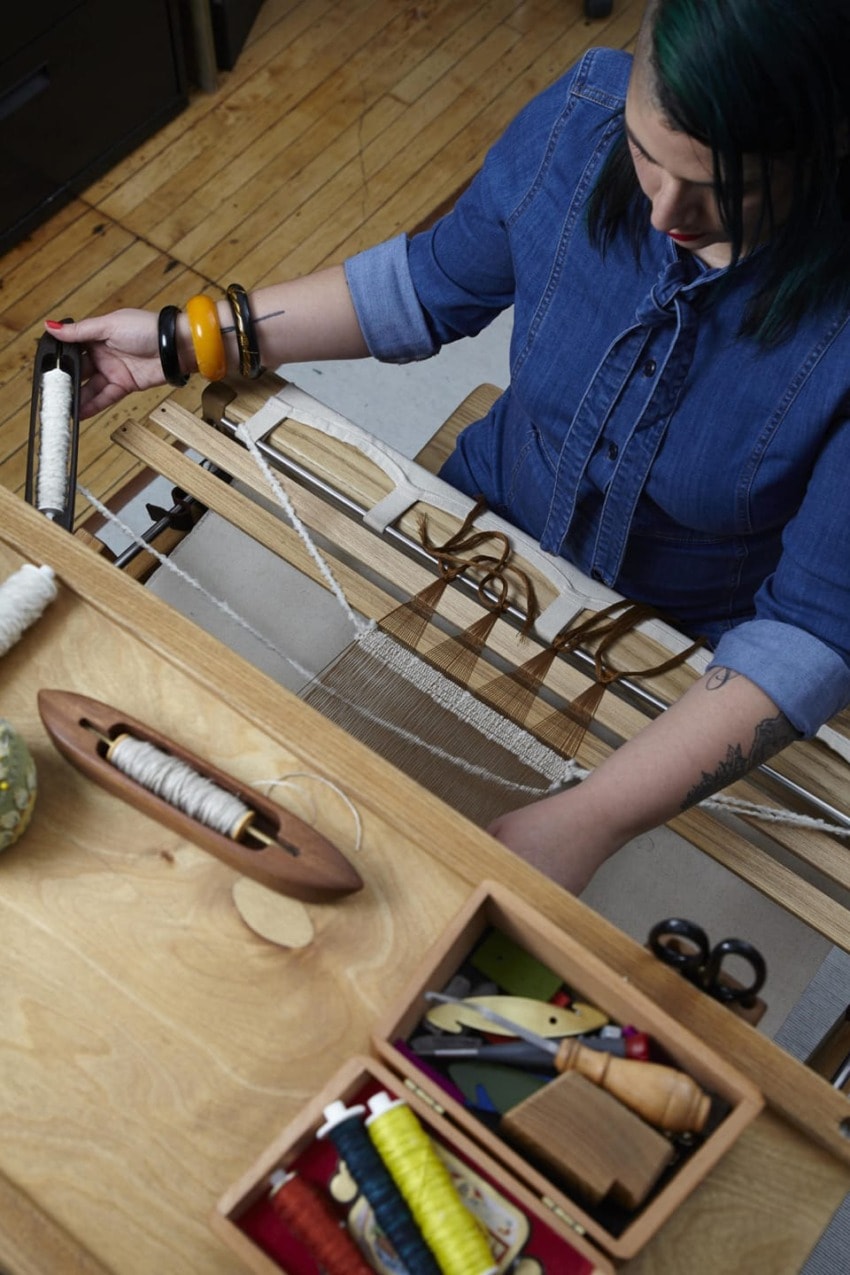 Person weaving on a loom with colorful threads and tools on a wooden desk in a cozy workspace.