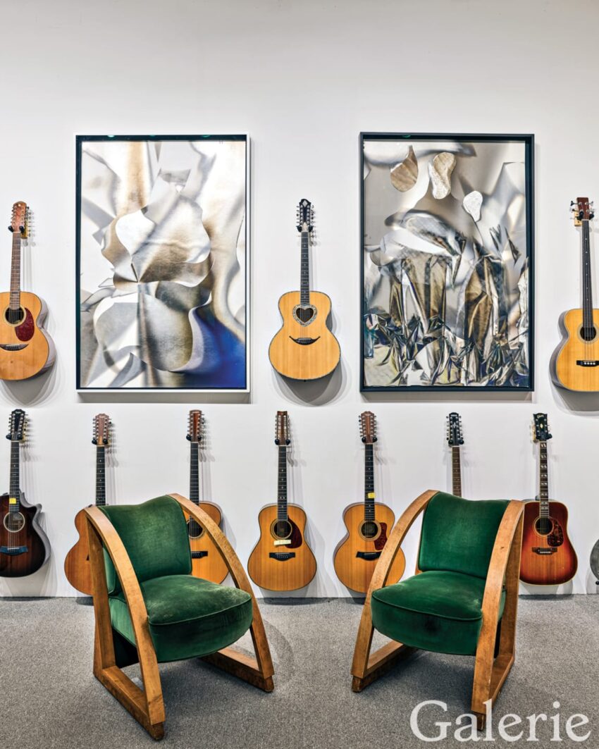 Gallery display with acoustic guitars on white walls, abstract art, and two green velvet chairs with wooden frames.