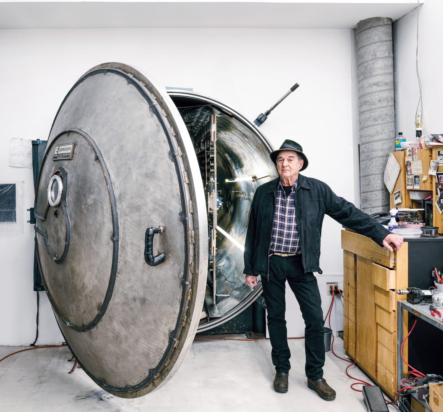 Man standing beside a large metal vault in an industrial setting, wearing a hat and plaid shirt, with tools and equipment nearby.