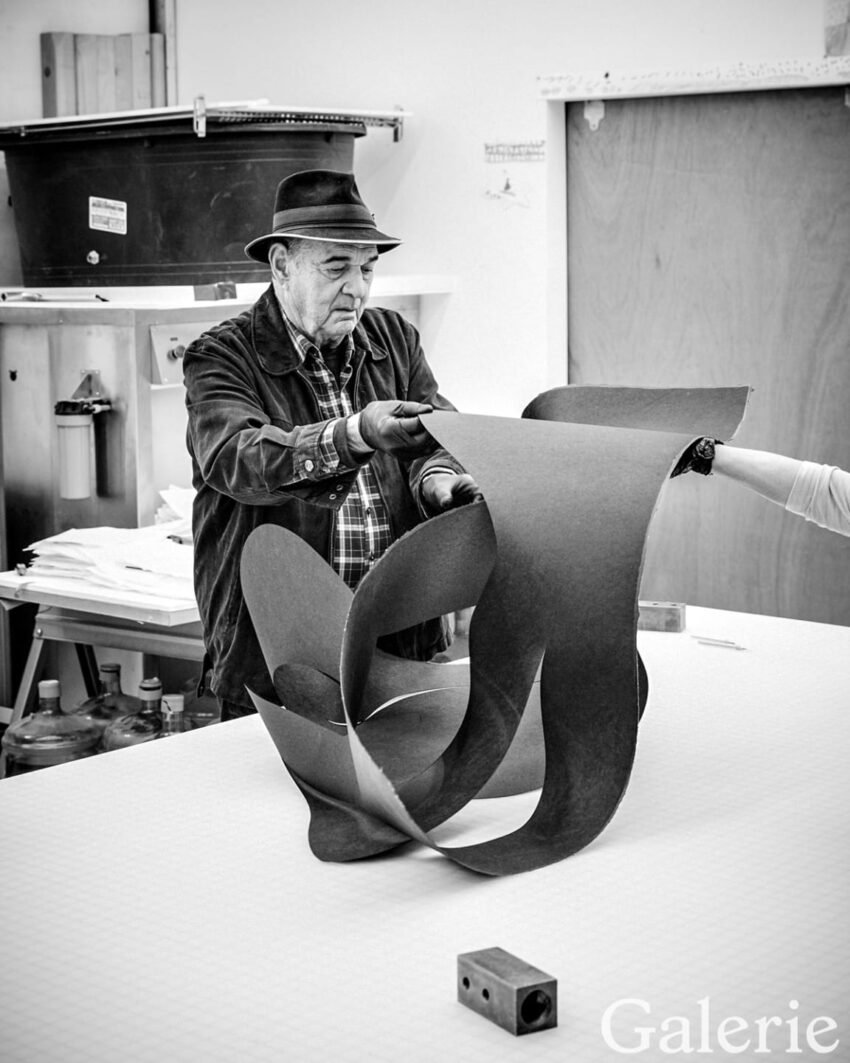 Man in hat shaping a large sheet in an art studio, with tools and materials on the table. Black and white image.