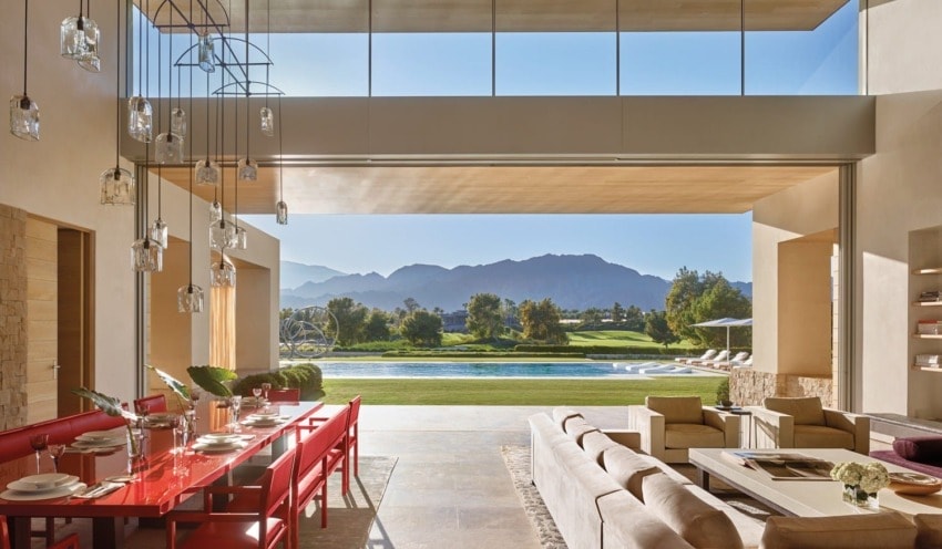 Open-air living room with red dining set, modern sofas, and a view of lush garden and mountains through large windows.
