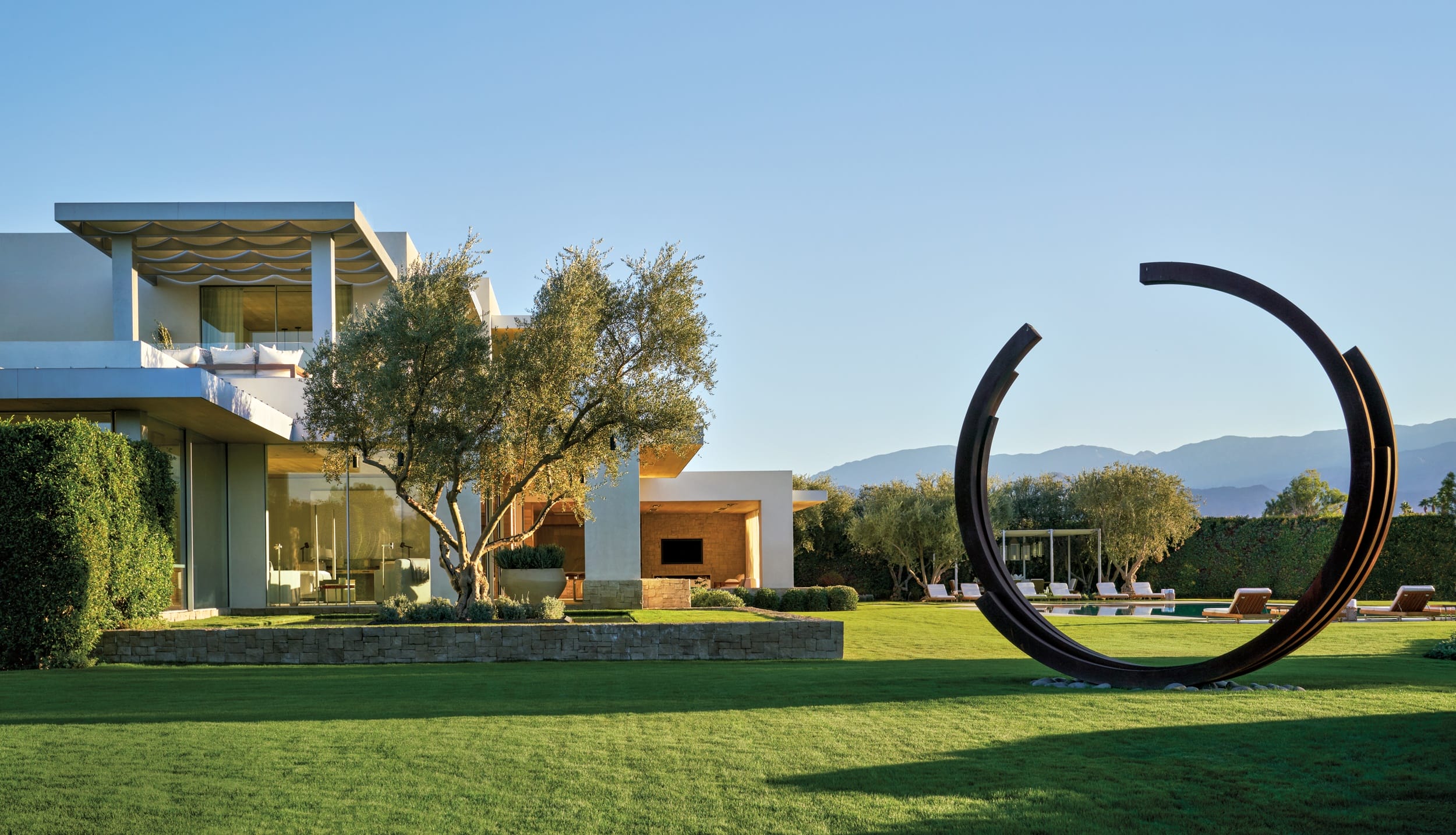 Modern house with large windows, manicured lawn, two trees, and circular sculpture on a sunny day.