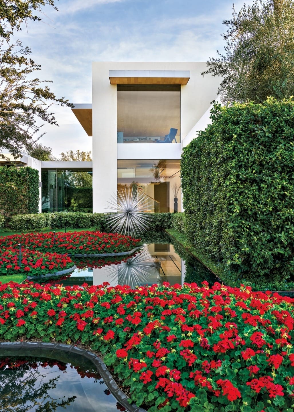 Modern house with large windows, surrounded by lush greenery and vibrant red flowers reflected in a serene pond.