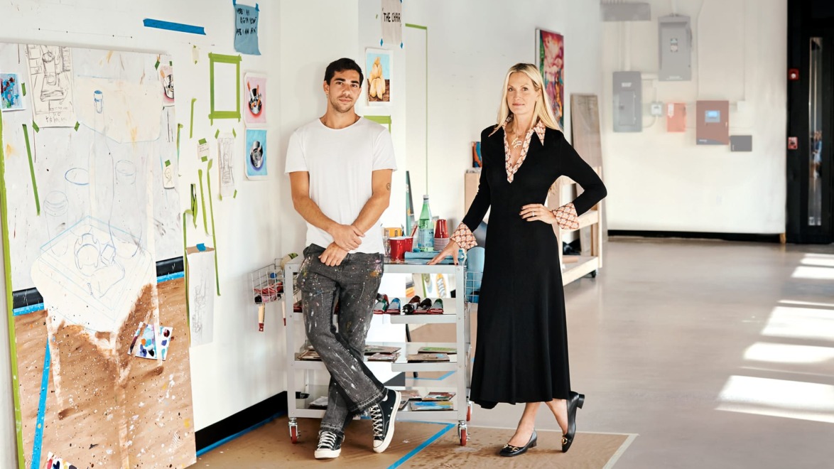 Two people standing in an art studio with paintings and art supplies in the background.