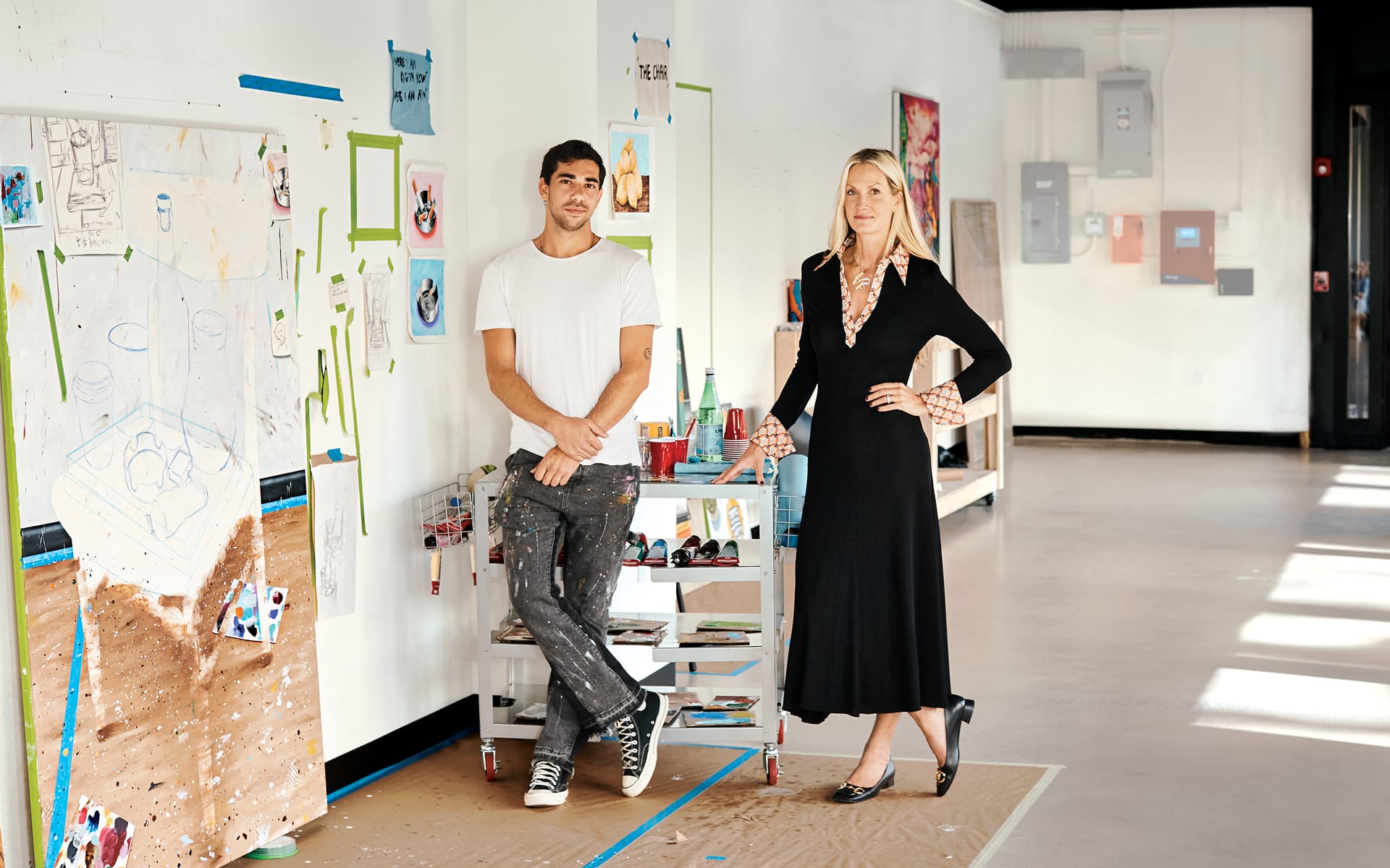 Two people standing in an art studio with paintings and art supplies in the background.