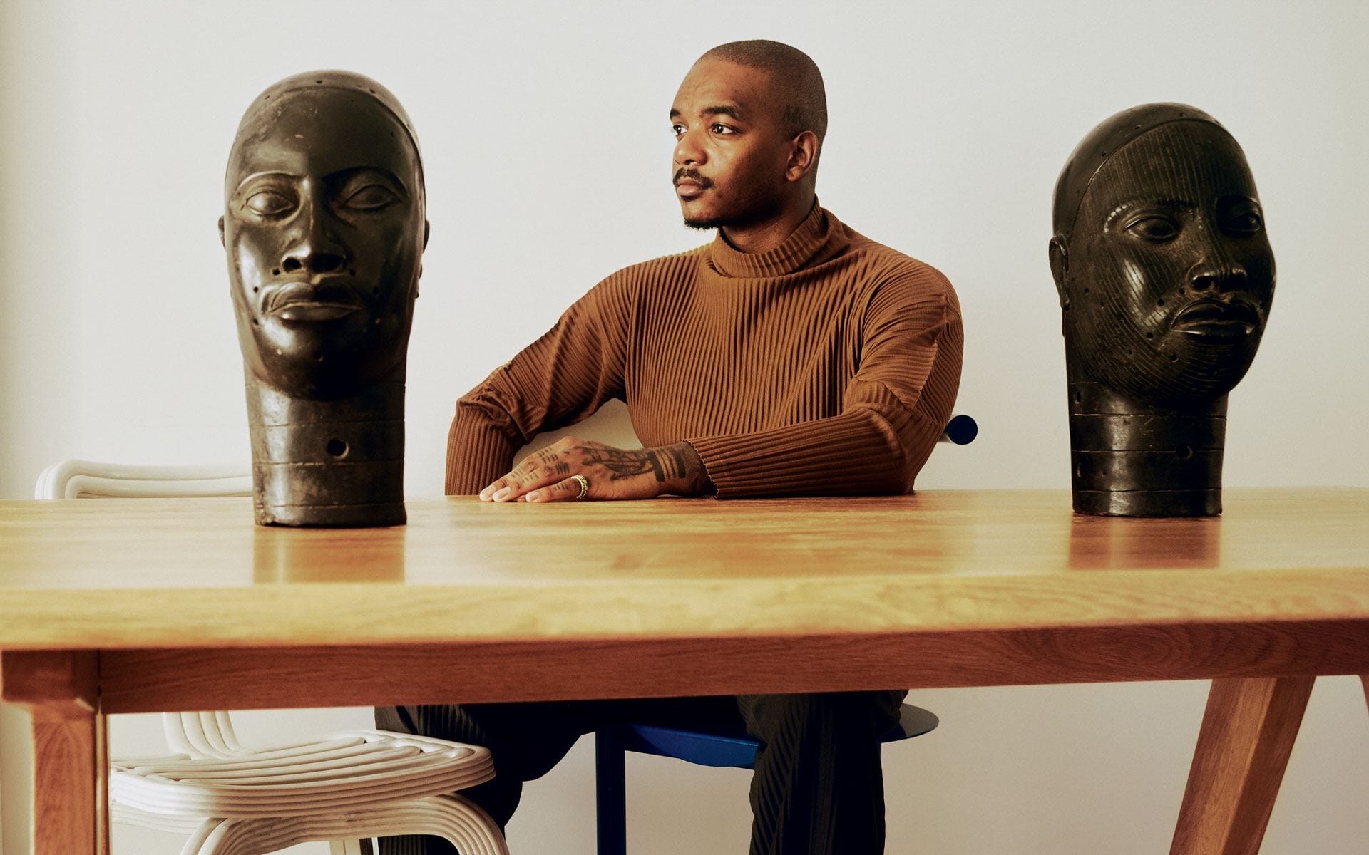 Man seated at a table with two sculpted busts, wearing a brown turtleneck, gazing thoughtfully to the side.