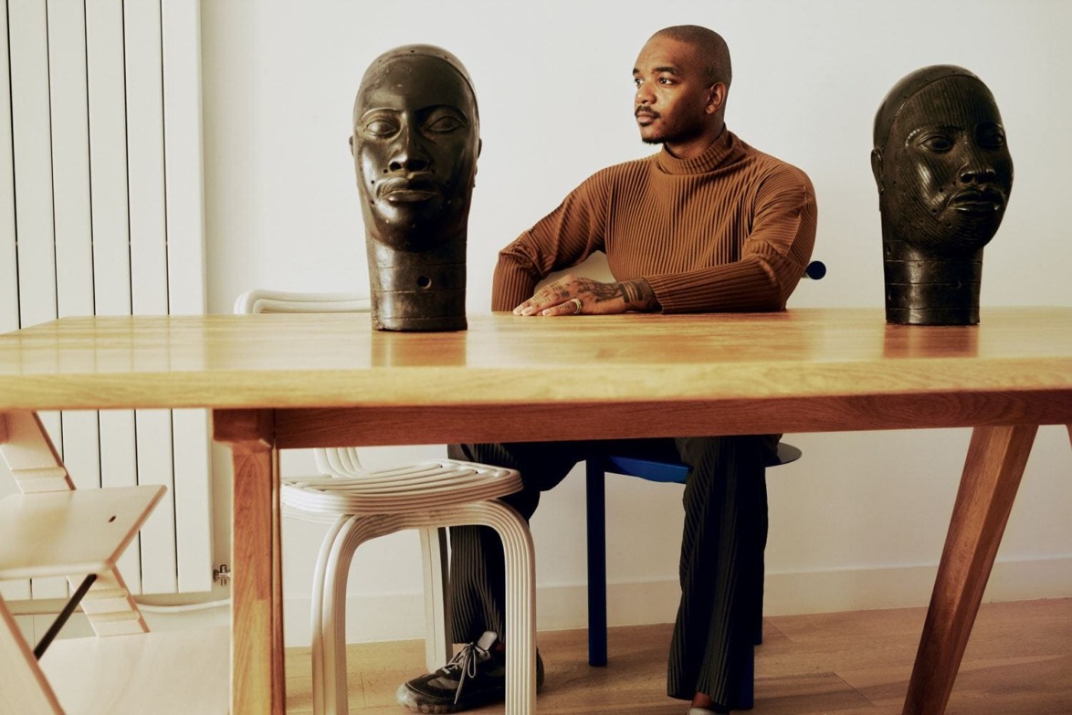 Man seated at a wooden table with two carved head sculptures, modern interior setting.