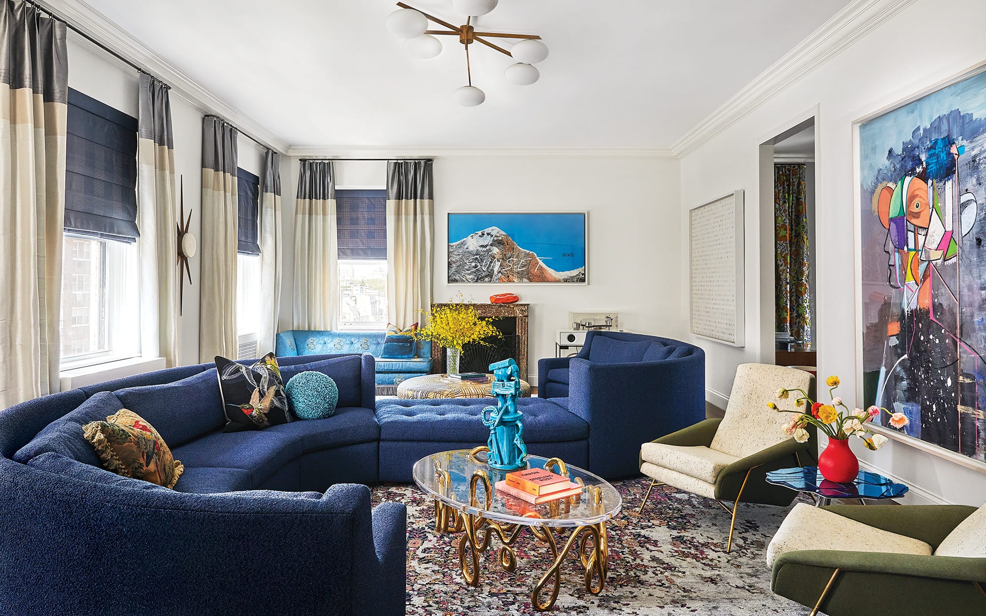 Modern living room with blue curved sofa, abstract wall art, and eclectic decor including vibrant sculptures and patterned rug.