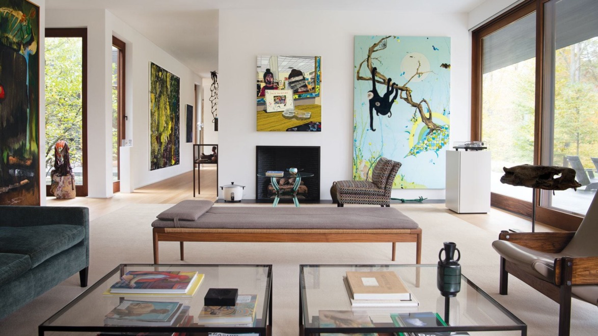 Modern living room with large windows, colorful abstract paintings, glass coffee tables, and wooden accents.