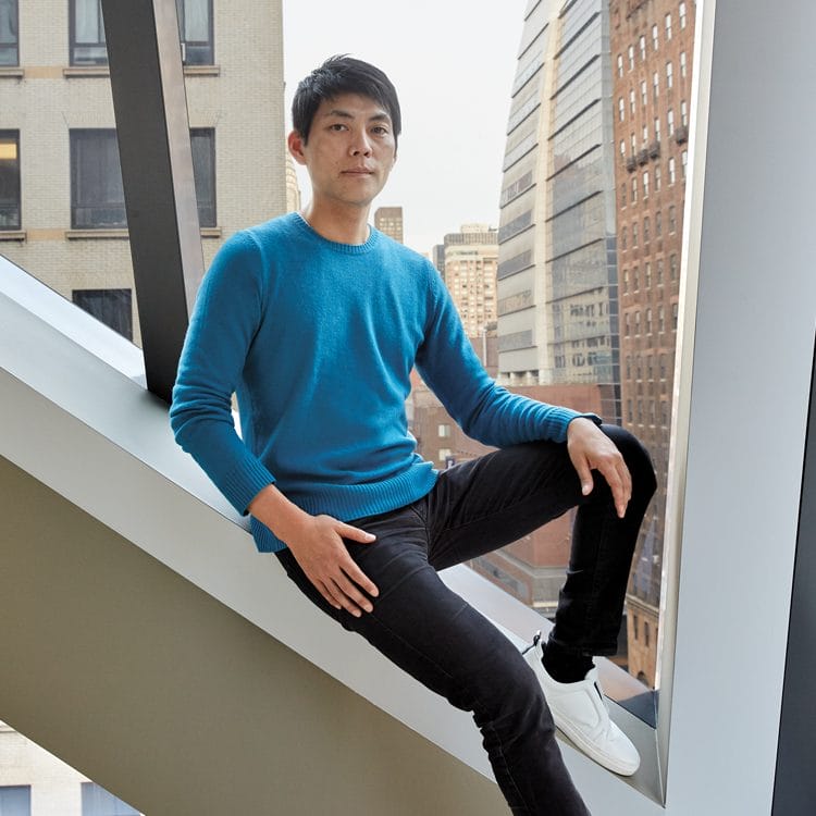 Person in a blue sweater and black pants sitting on a modern architectural feature with city buildings in the background.