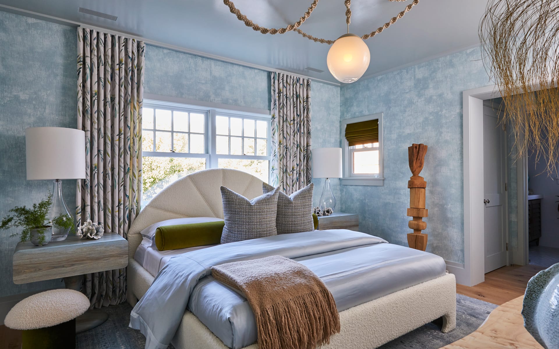 Modern bedroom with blue walls, patterned curtains, white bed, and unique light fixture. Wood accents and natural light.