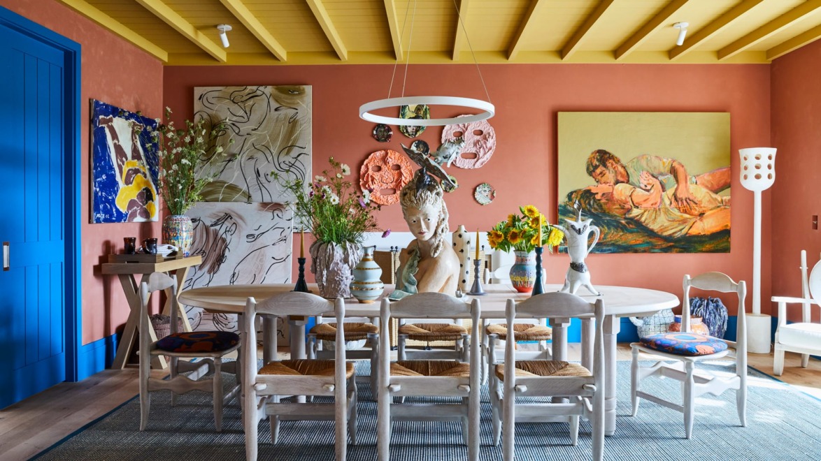 Eclectic dining room with colorful art, round table, white chairs, and vibrant decor under a yellow ceiling.