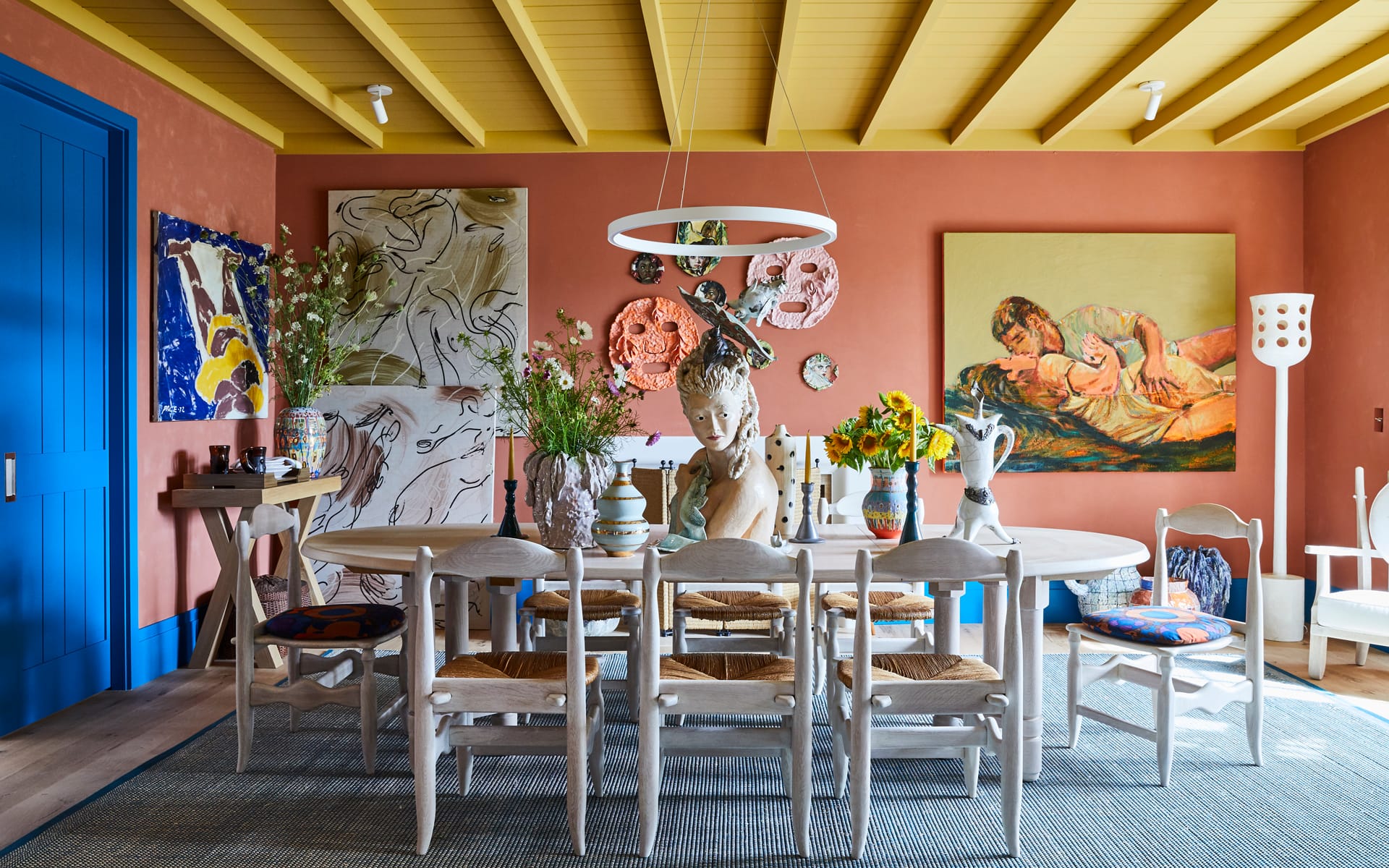 Eclectic dining room with colorful art, round table, white chairs, and vibrant decor under a yellow ceiling.