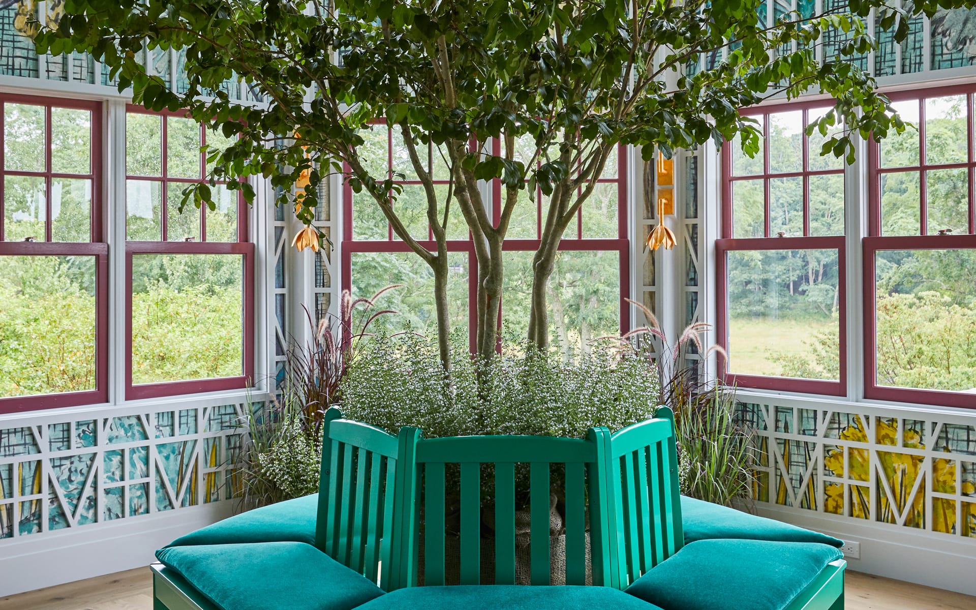 Tree grows through vibrant green bench in sunroom with large windows and patterned walls, overlooking lush greenery.