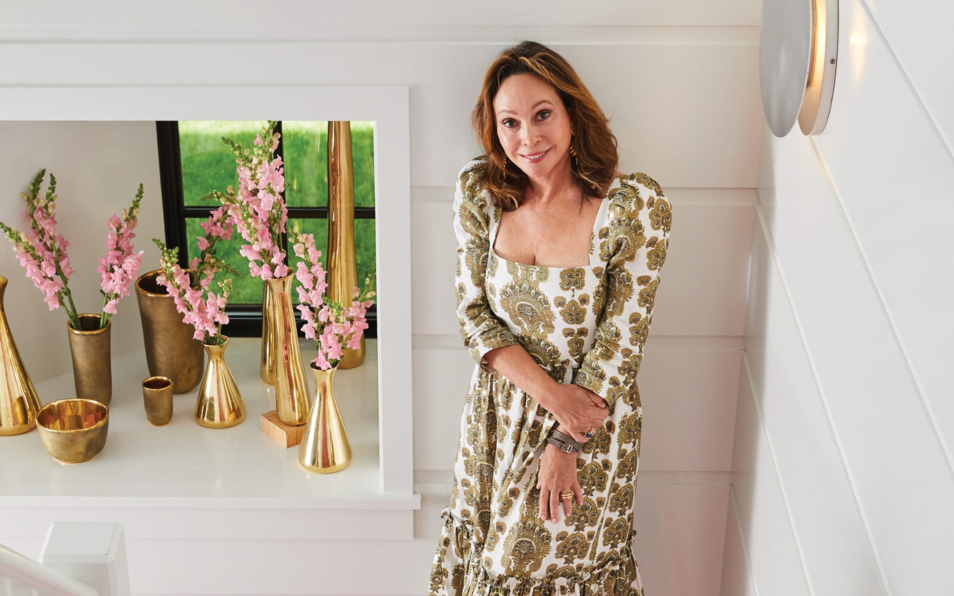 Woman in floral dress standing by window with pink flowers in gold vases, smiling in a white interior setting.