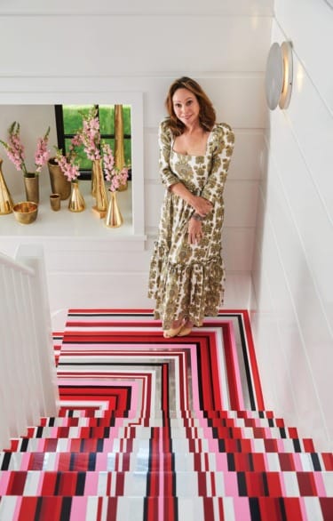Woman in a patterned dress stands on vibrantly striped staircase with floral vases in the background.