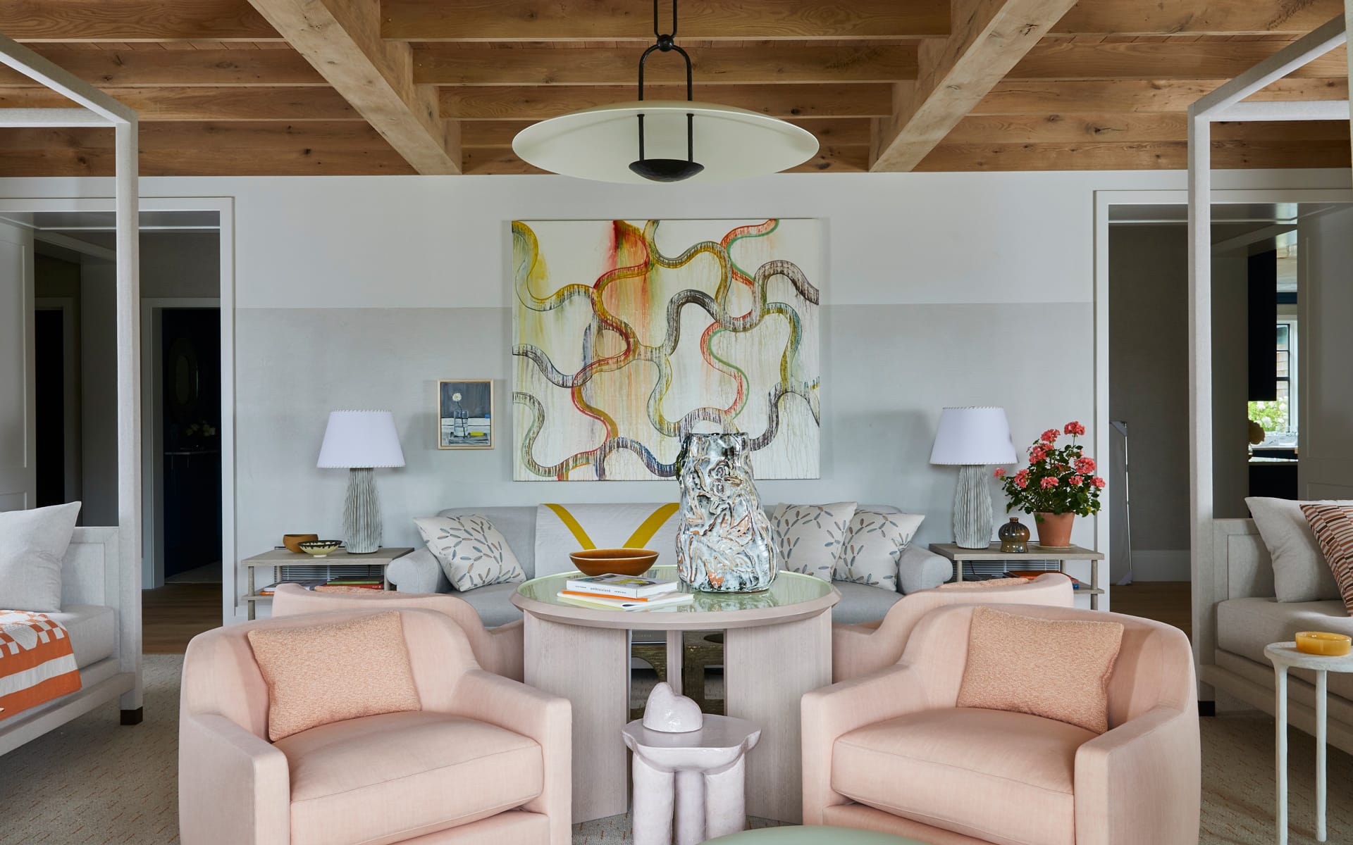 Modern living room with abstract art, two pink chairs, a white sofa, wooden ceiling beams, and decorative vases on a table.