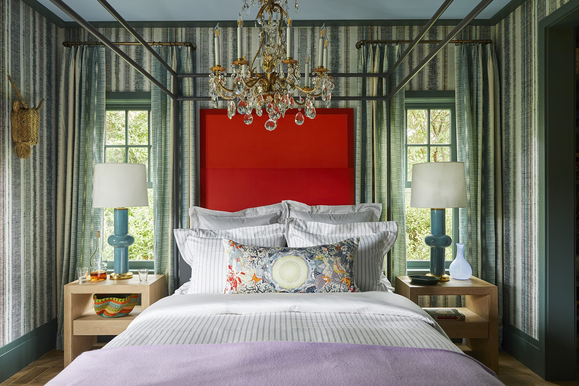 Eclectic bedroom with a four-poster bed, red headboard, striped walls, and a crystal chandelier, featuring colorful decor accents.