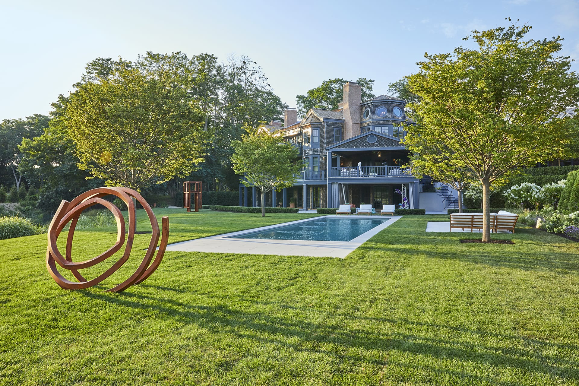 Spacious backyard with a modern sculpture, swimming pool, and large house in the background surrounded by trees.
