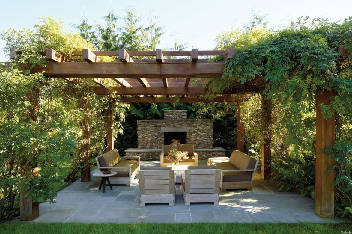 Outdoor pergola with wooden furniture and fireplace surrounded by lush greenery in a garden setting.