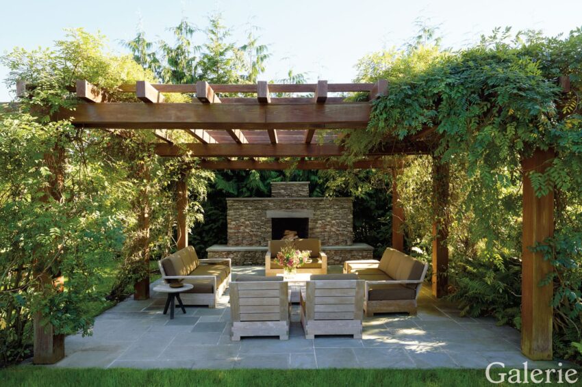 Outdoor patio with wooden pergola, stone fireplace, surrounding greenery, and comfortable seating for relaxation.