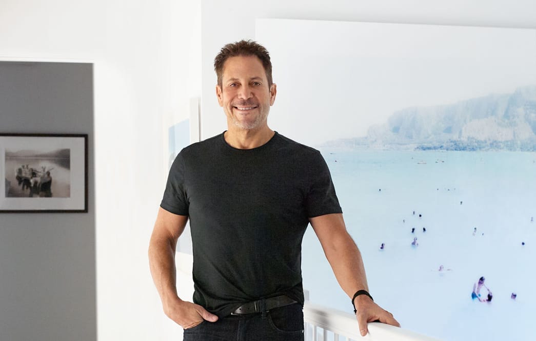 Man standing in a modern room with framed art on the wall and wearing a black t-shirt and watch.