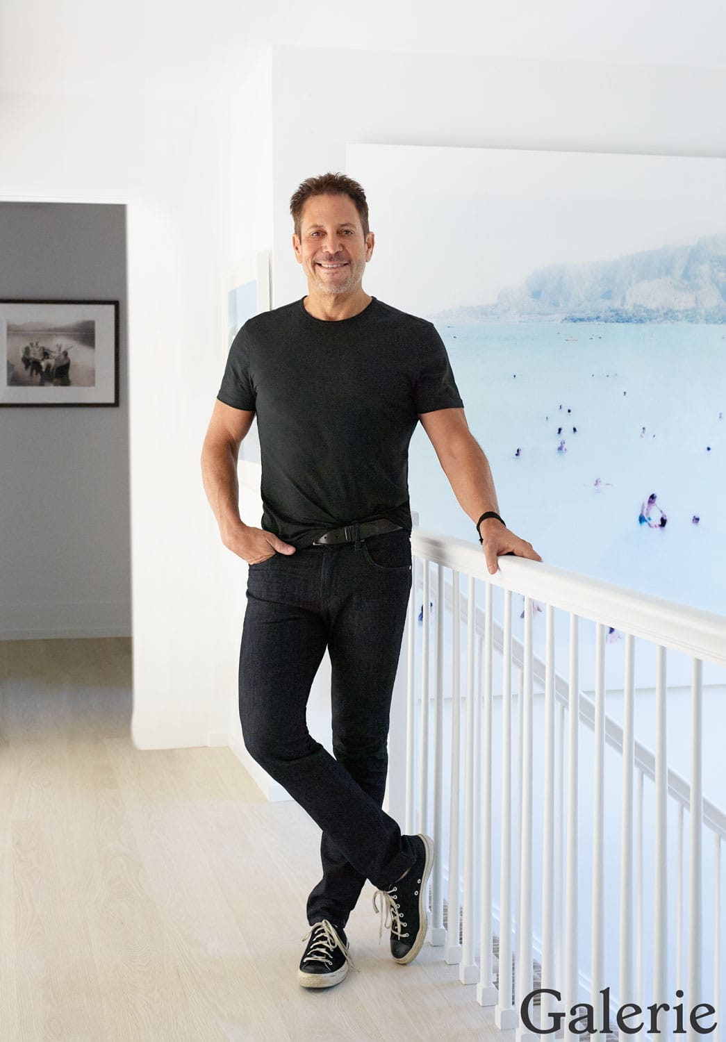 Man in black t-shirt and jeans leaning on a white railing in a bright, modern interior setting.