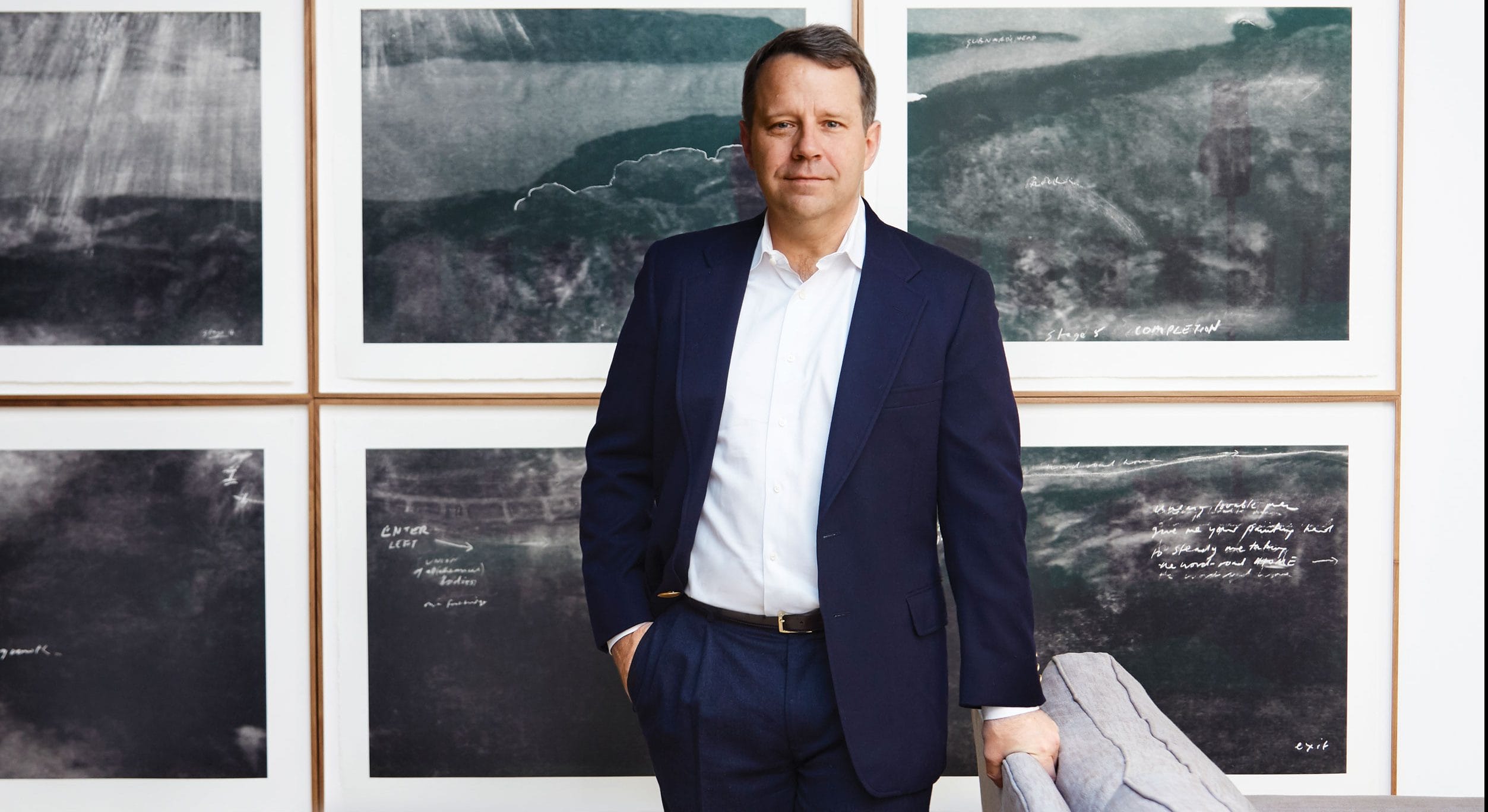 Man in a suit standing in front of framed abstract artwork with a serious expression.