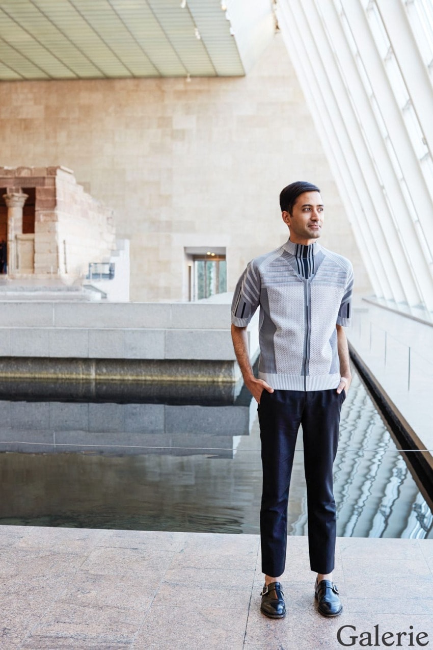 Person standing in a modern gallery by a reflective indoor pool, dressed in stylish clothing, with large windows in the background.