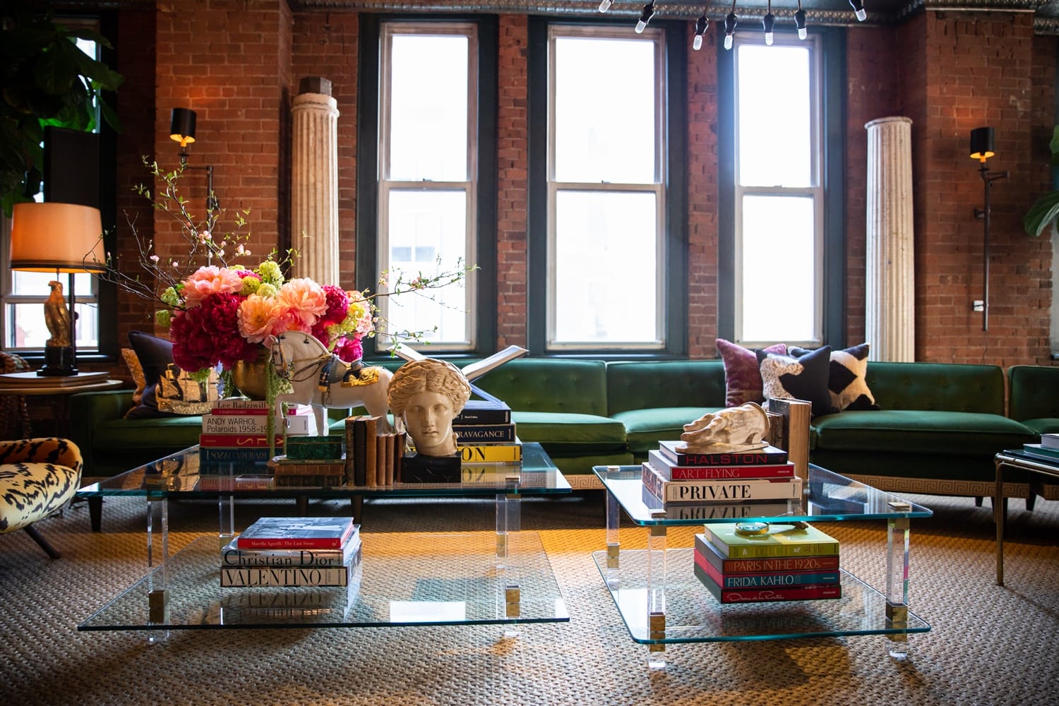 Chic living room with glass tables, books, colorful flowers, and a green velvet sofa against exposed brick walls.