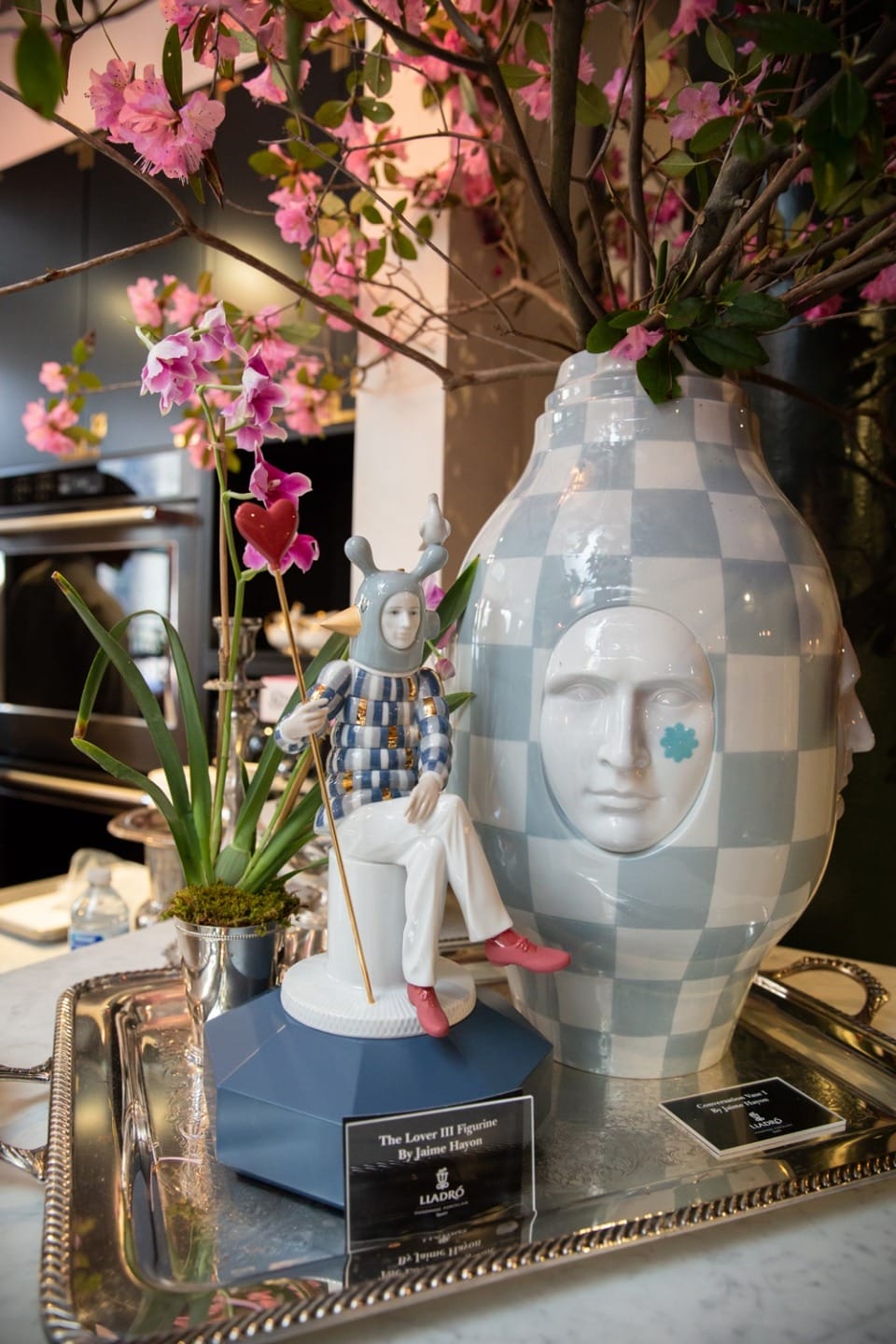 Porcelain figure on tray with large vase and pink orchids in a modern kitchen setting.