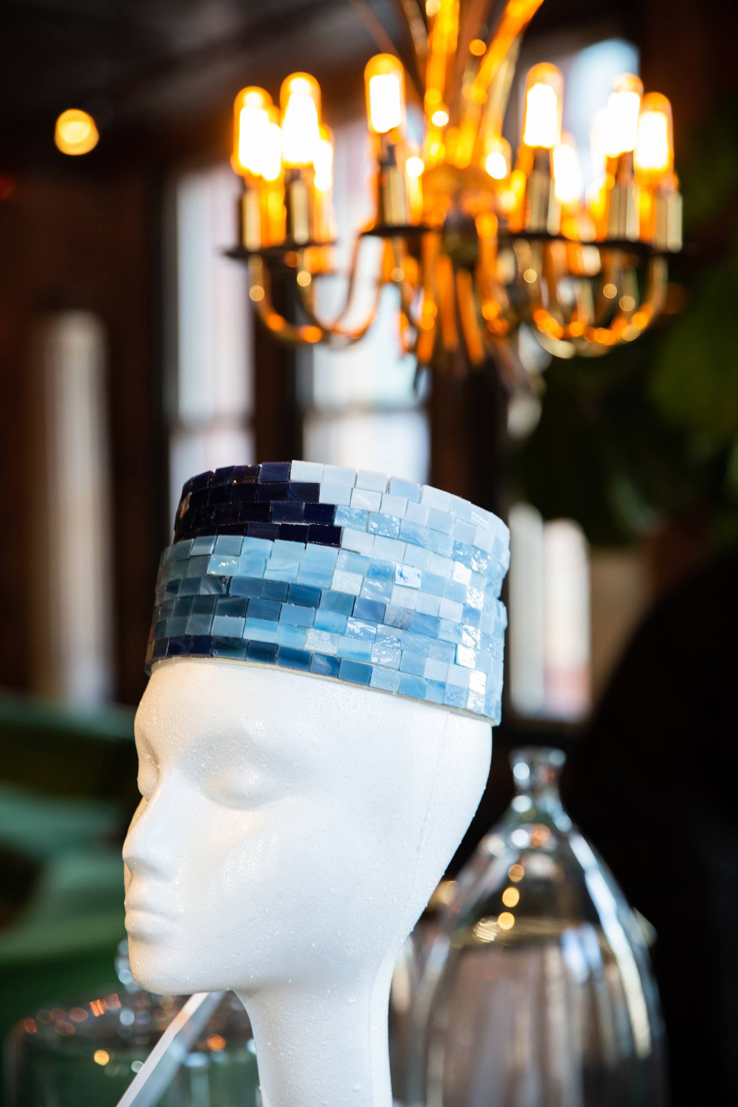 Styrofoam mannequin with a mosaic tile hat under a lit chandelier in a stylish room.