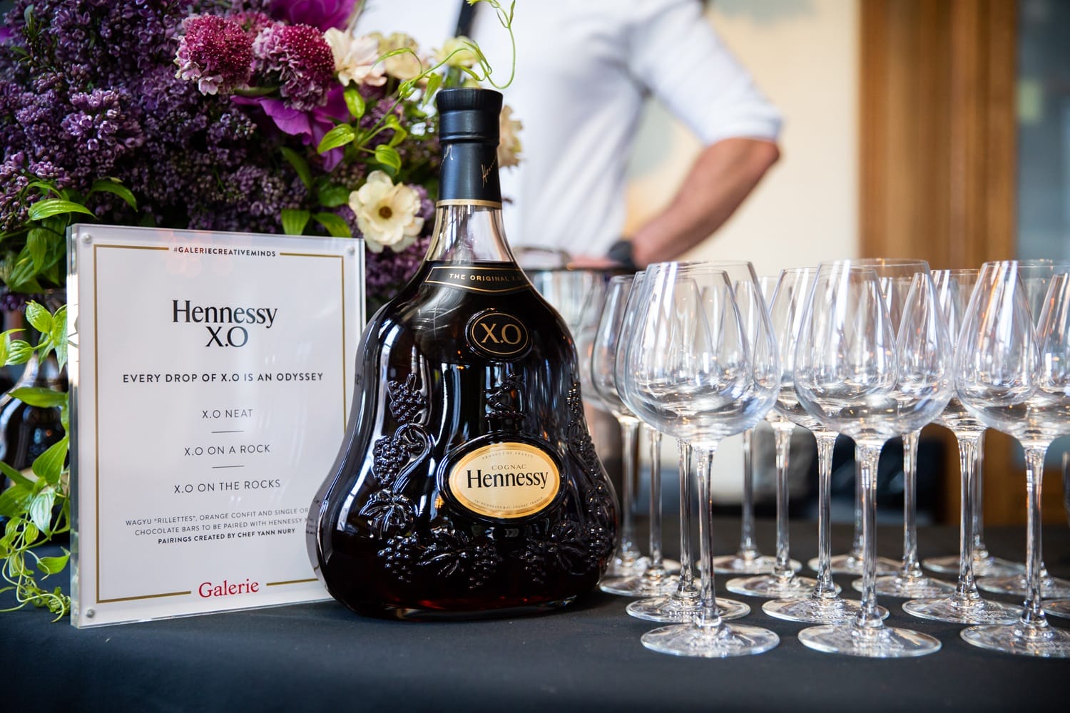 Hennessy XO bottle on a table with a display sign and several empty wine glasses arranged nearby.