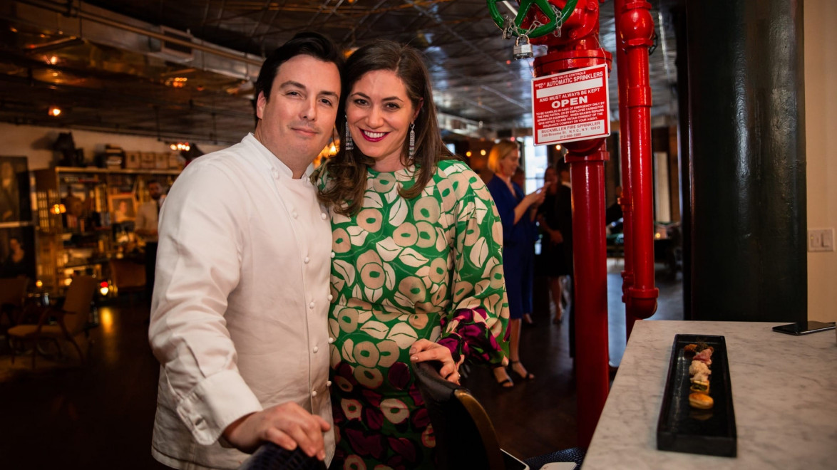 Chef in white coat and smiling woman in patterned dress stand together in a warmly lit restaurant near a red valve.