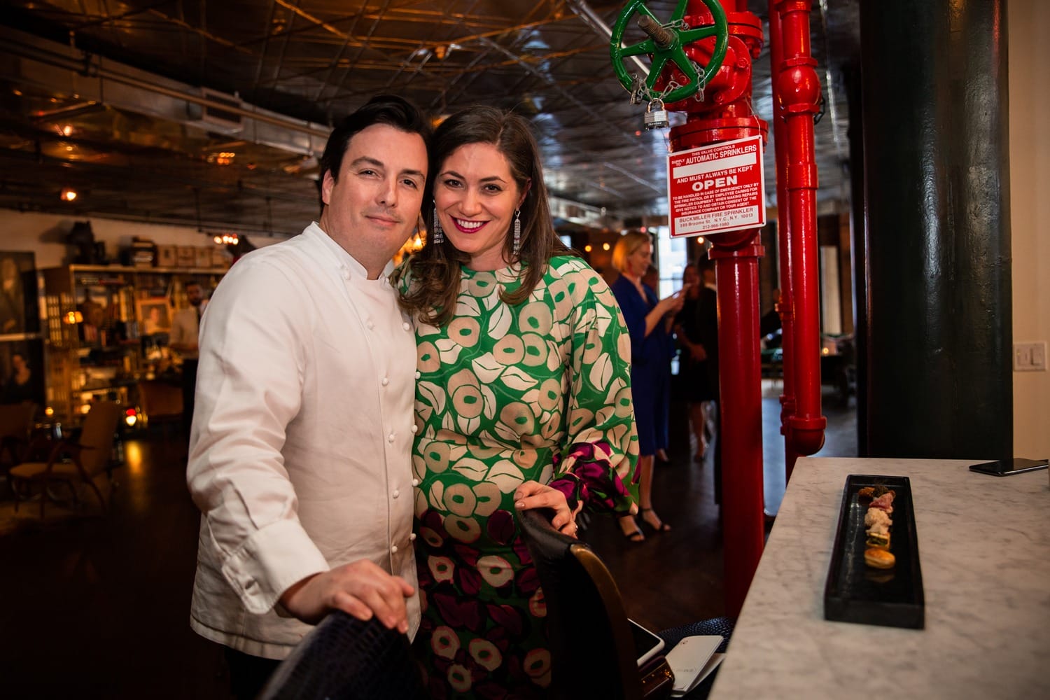 A couple poses together in a warmly lit restaurant, with a chef wearing white and a woman in a colorful dress.