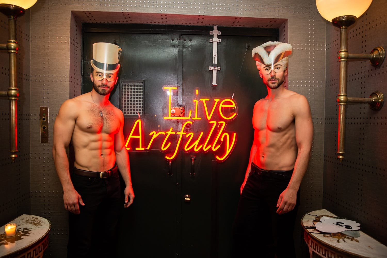 Two shirtless men with hats standing next to a neon sign reading "Live Artfully" in a dimly lit room.