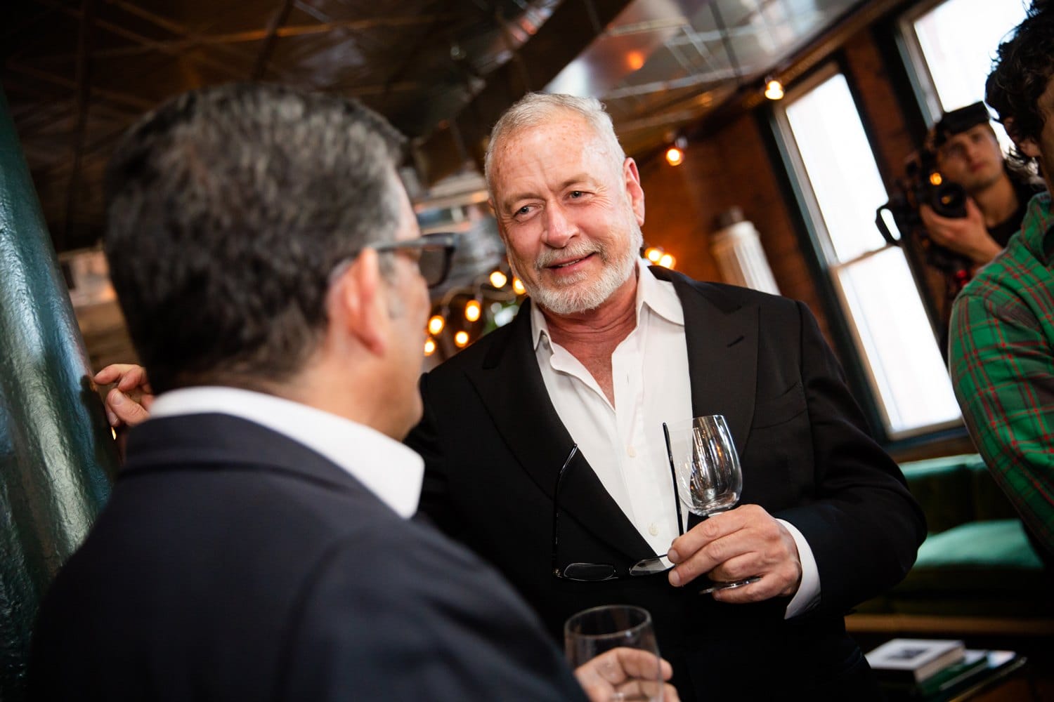 Two men in a lively room, dressed in suits, engaged in conversation while holding wine glasses.