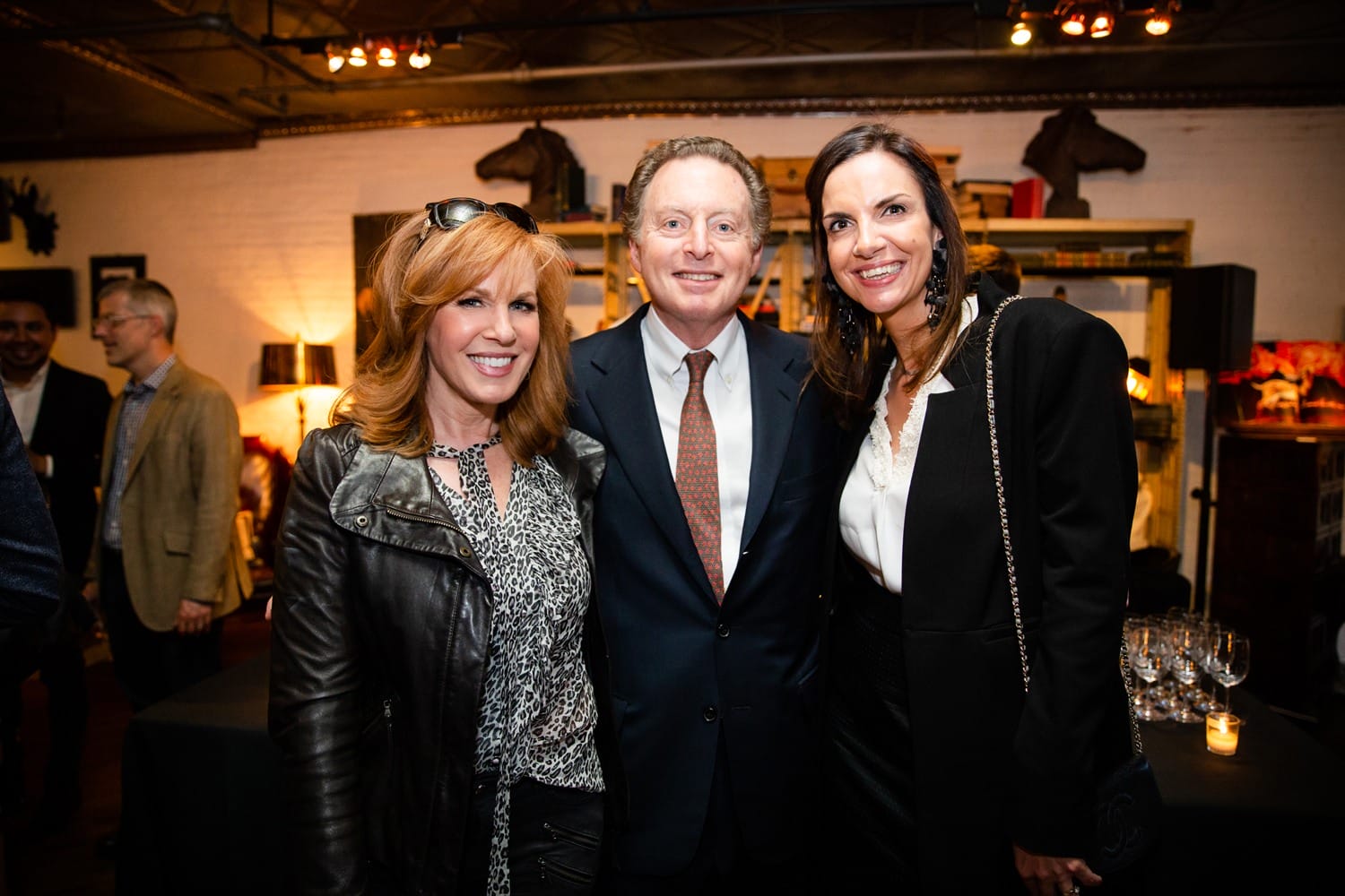 Three people smiling and posing in a warmly lit room with decorative shelves and art in the background.