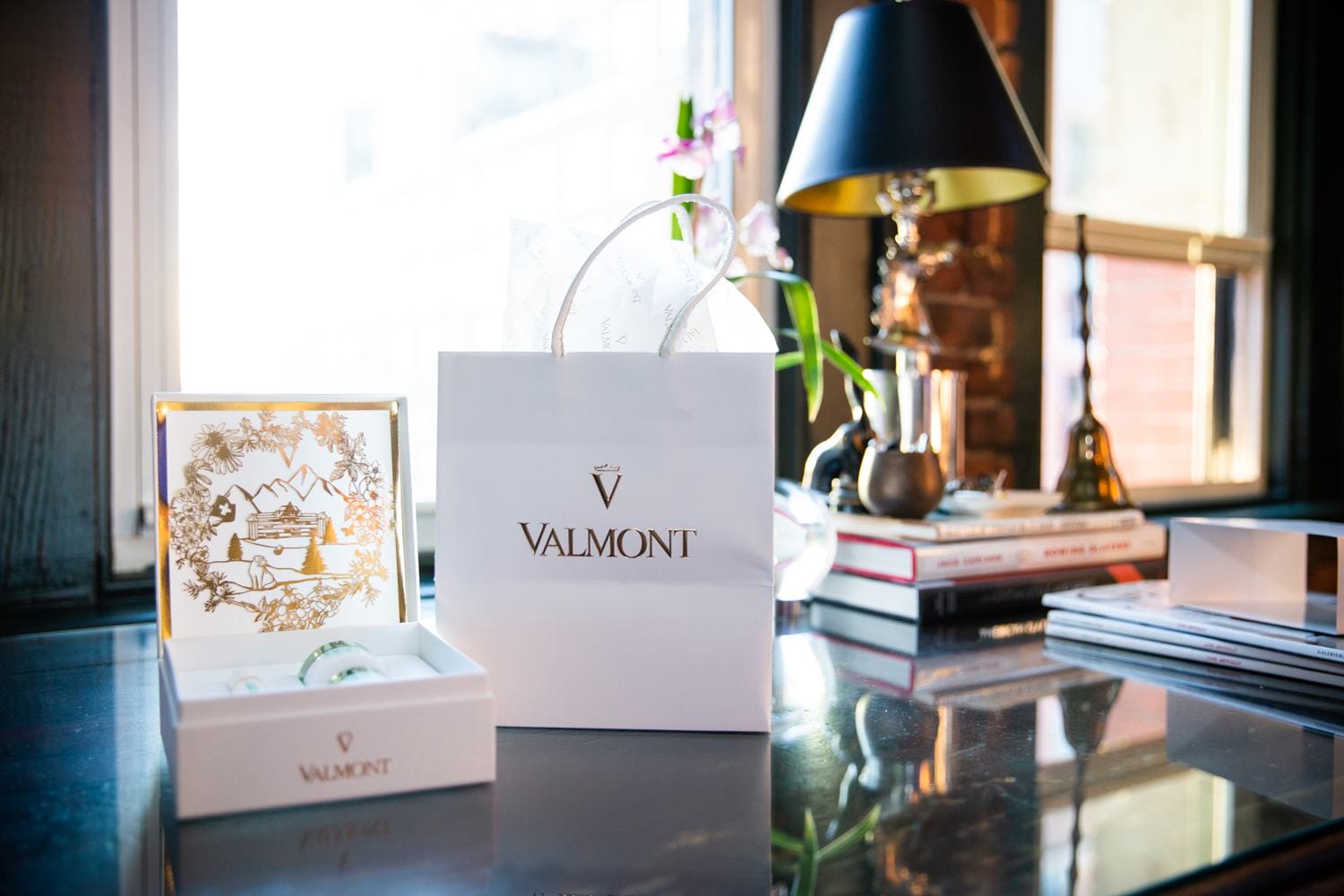 Valmont gift bag and open box with decorative design on a reflective table near a window and lamp.