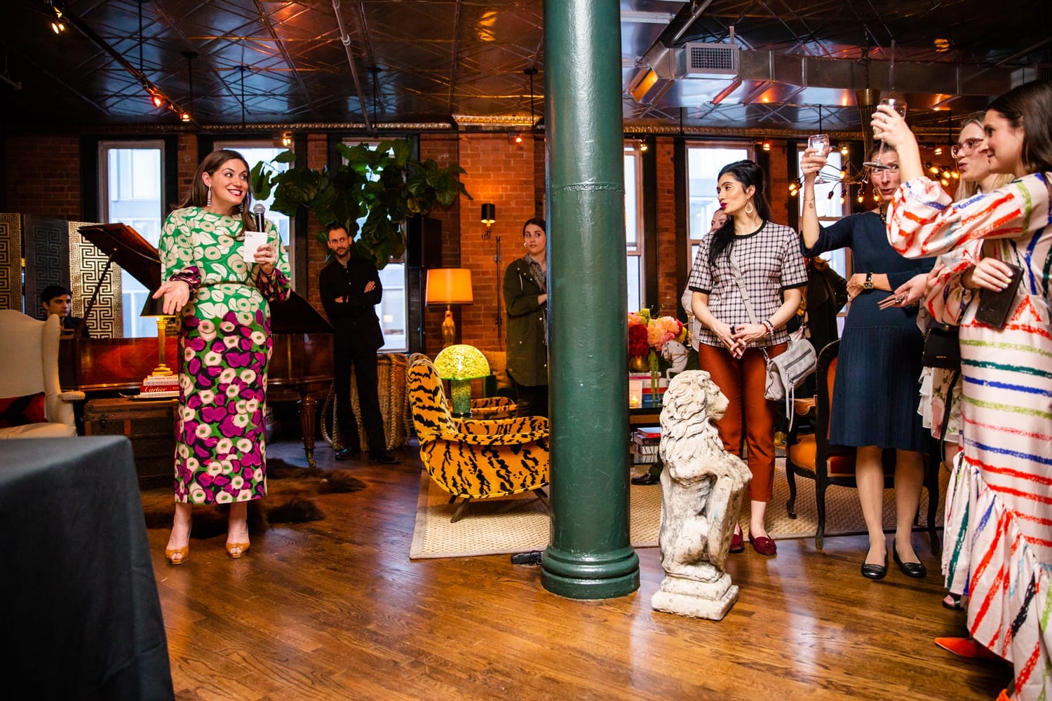 Group of people socializing in a warmly lit, stylish room with colorful attire and a woman giving a toast.