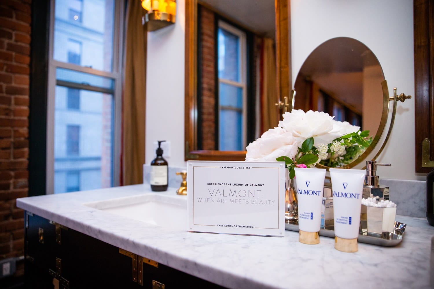 Luxurious bathroom countertop with Valmont beauty products, white flowers, and a mirror reflecting city view.